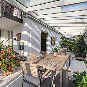 Moderne überdachte Terrasse mit Holztisch, Gartenmöbeln und vielen grünen Pflanzen.