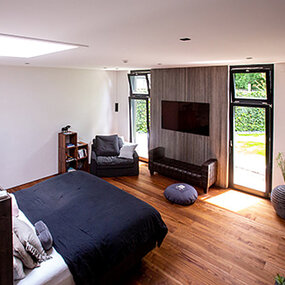 Modernes Schlafzimmer mit Holzboden, großen Fenstern, Doppelbett und gemütlicher Sitzecke.